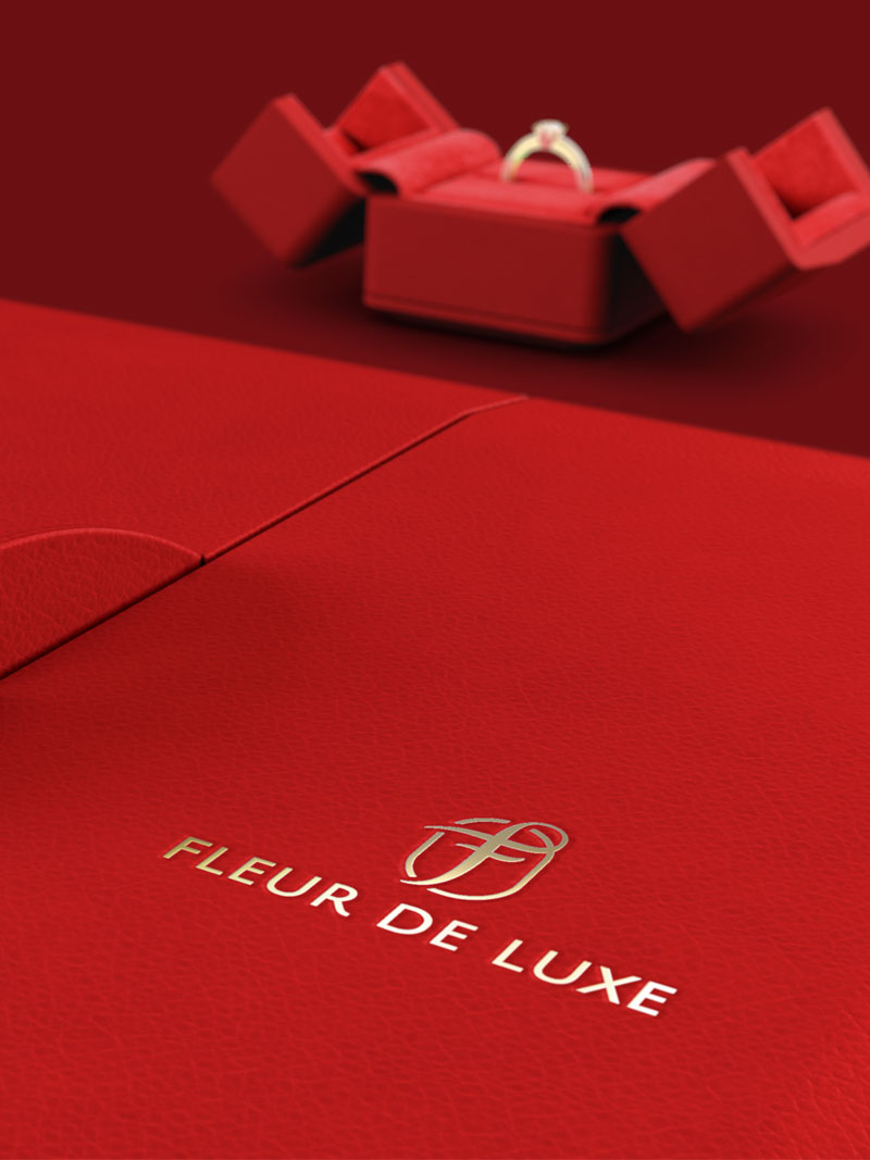 Détail d'un écrin à collier Fleur de Luxe rouge avec logo estampé à la feuille d'or, en arrière-plan étui à anneaux rouge ouvert