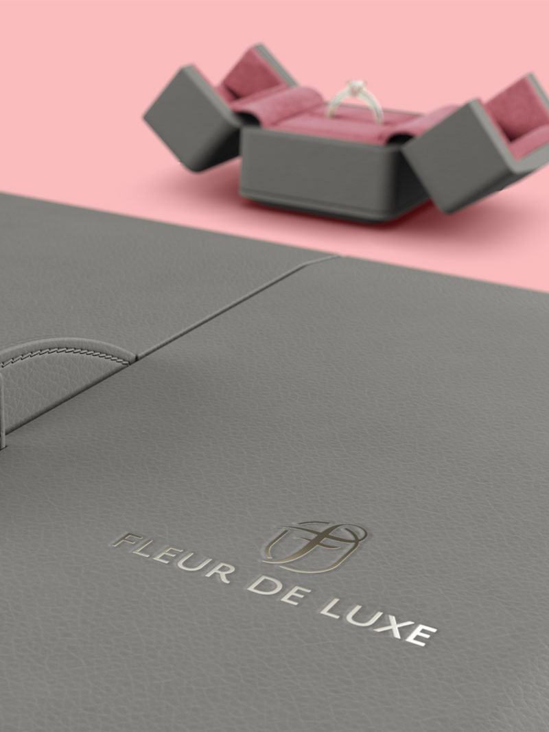 Détail de l'écrin à collier Fleur de Luxe en gris-brun avec le logo en feuille d'argent, en arrière-plan un étui à anneaux ouvert.